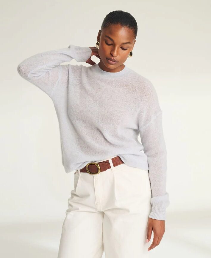 A person wearing a light gray sweater and white pants with a brown belt stands against a plain background, resting one hand on the back of their neck.