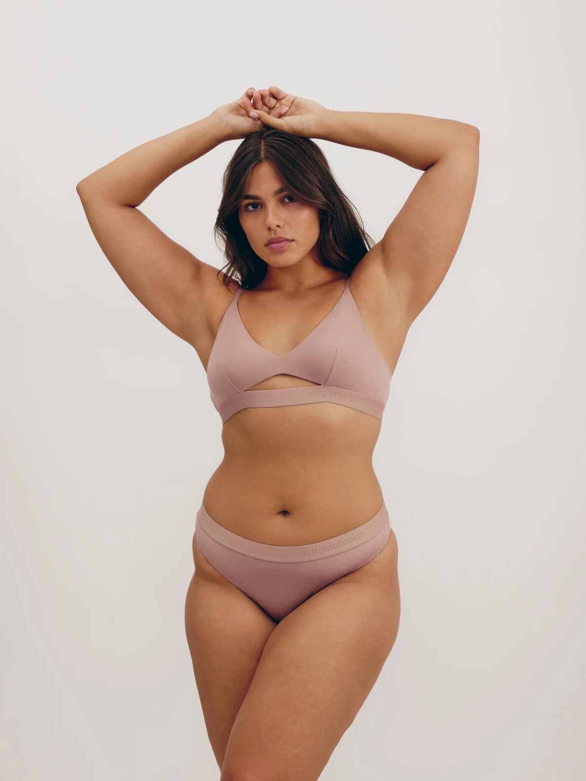 A woman stands against a plain background wearing a matching light mauve bra and underwear set, with her arms raised above her head.