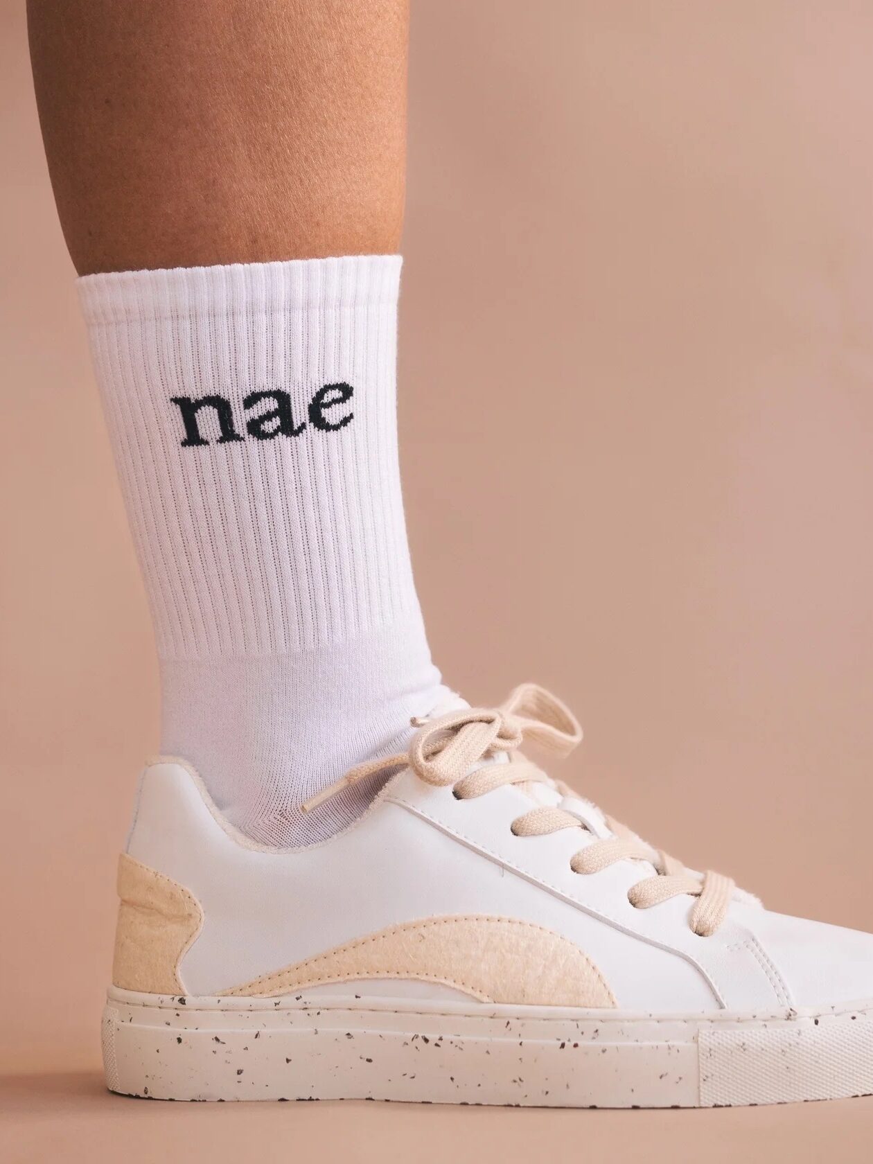 A person wearing a white sneaker and a white ribbed sock with the word "nae" printed on it, standing against a beige background.