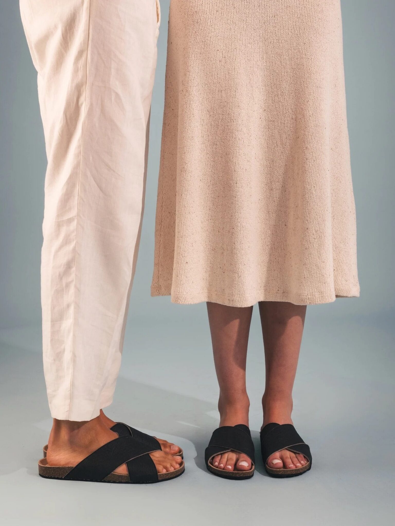 Two people standing side by side wearing light-colored clothing and black sandals, with only their legs and feet visible against a plain background.