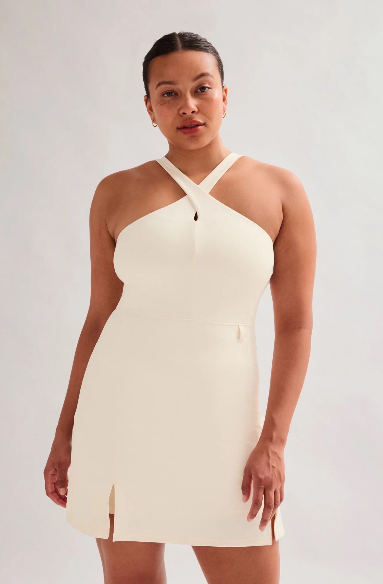 A woman stands against a plain background wearing a sleeveless, cream-colored dress with a cross halter neckline and small front slits.