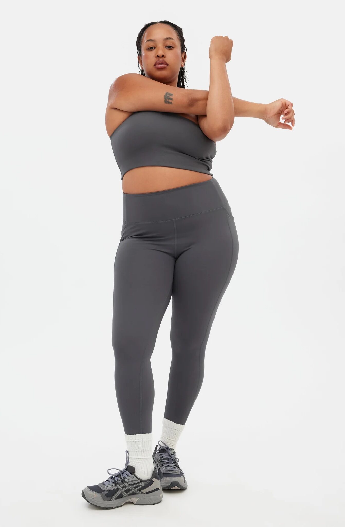 Person wearing a grey sports bra and leggings with white socks and grey sneakers, standing and stretching one arm across their chest against a plain background.