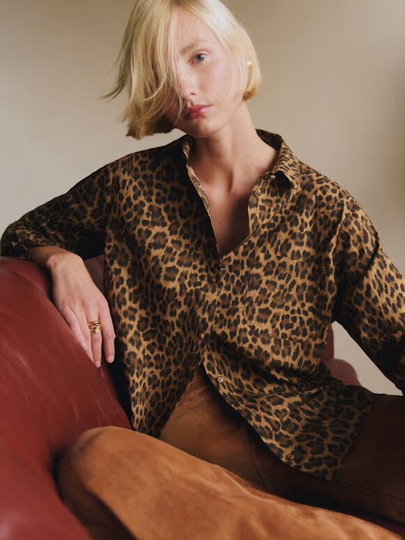 A person with short blonde hair sits on a red-brown couch, wearing a leopard-print shirt and brown pants, looking at the camera.