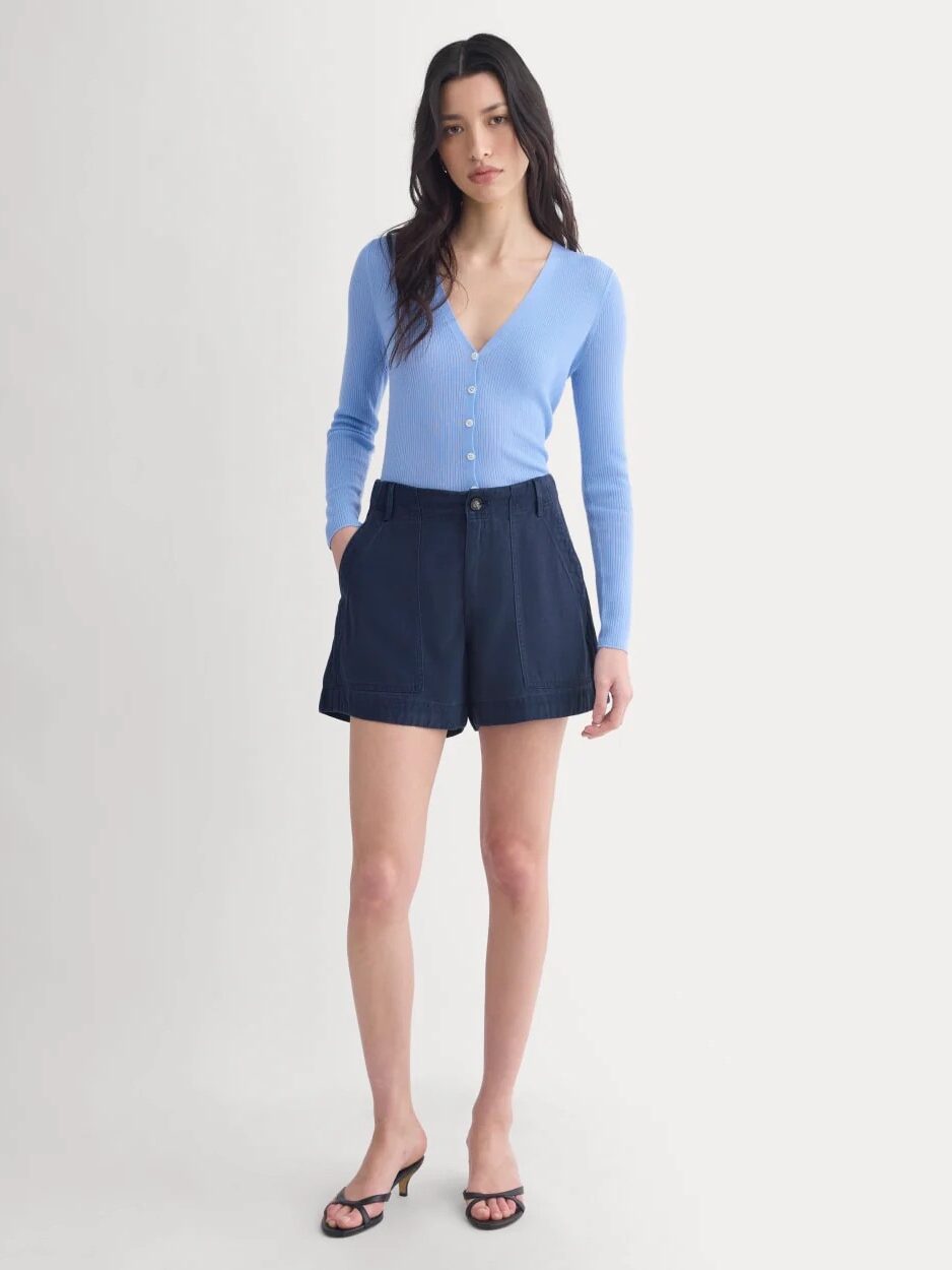 Woman standing against a plain background, wearing a light blue buttoned long-sleeve top, navy high-waisted shorts, and black sandals. Hands in pockets, facing forward.