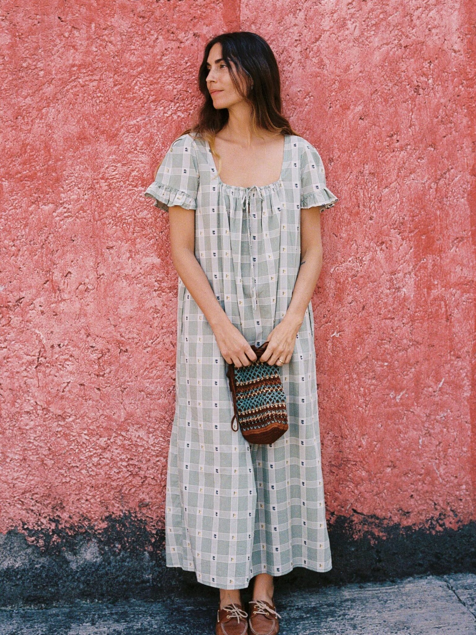 A woman in a long, patterned dress stands against a textured pink wall, holding a small woven bag and looking to the side.