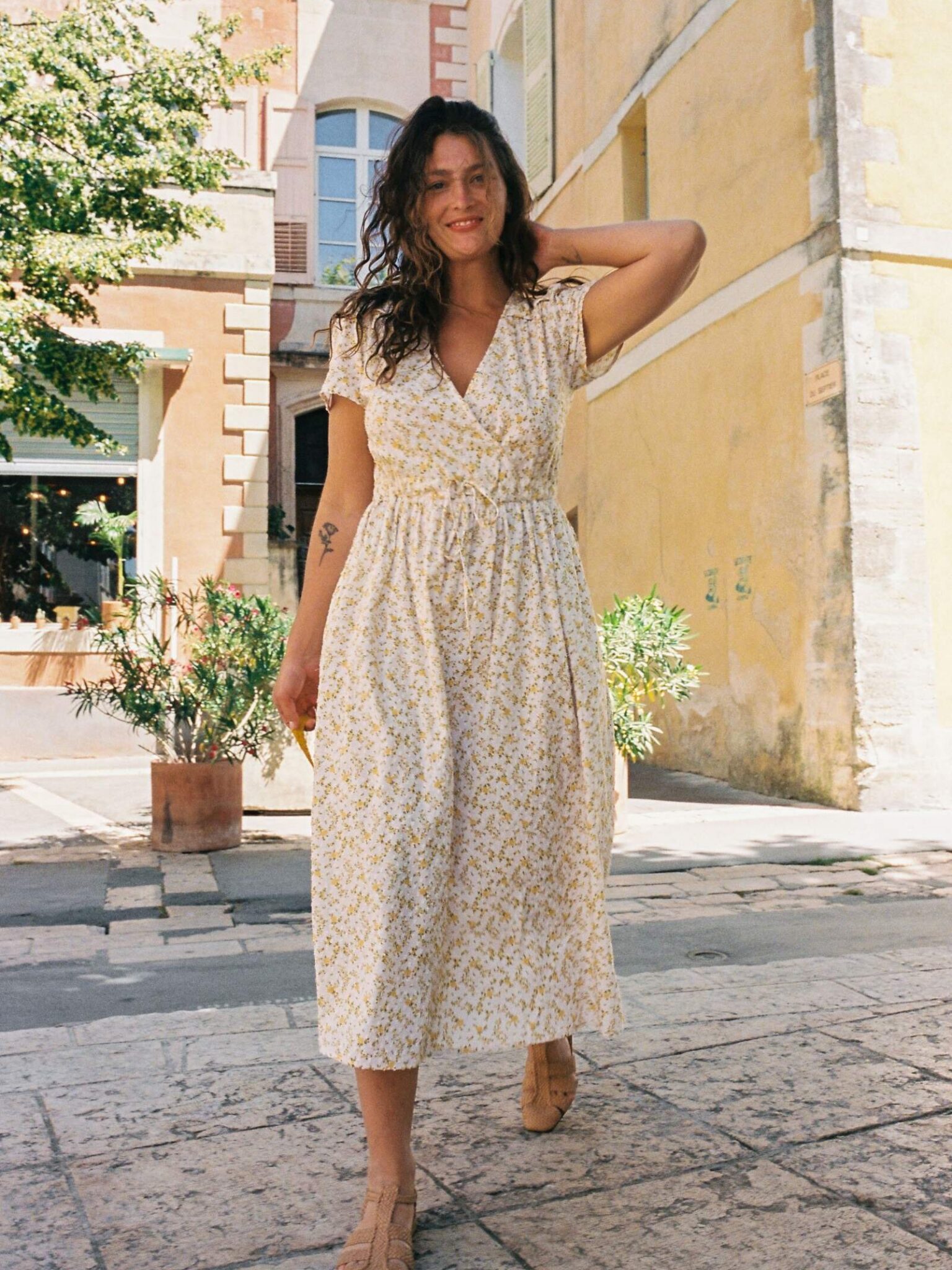 A woman in a light floral dress and tan shoes stands outdoors on a sunny day, with buildings and potted plants in the background.