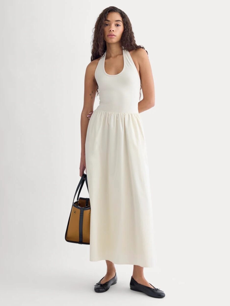 A woman wearing a sleeveless, halter-neck cream dress with a gathered waist, black flats, and holding a tan handbag stands against a plain light background.