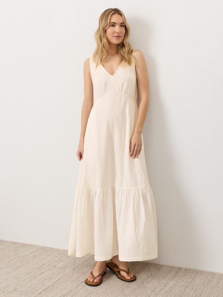 A woman stands against a white wall wearing a sleeveless, long cream-colored dress and brown sandals on a light textured floor.