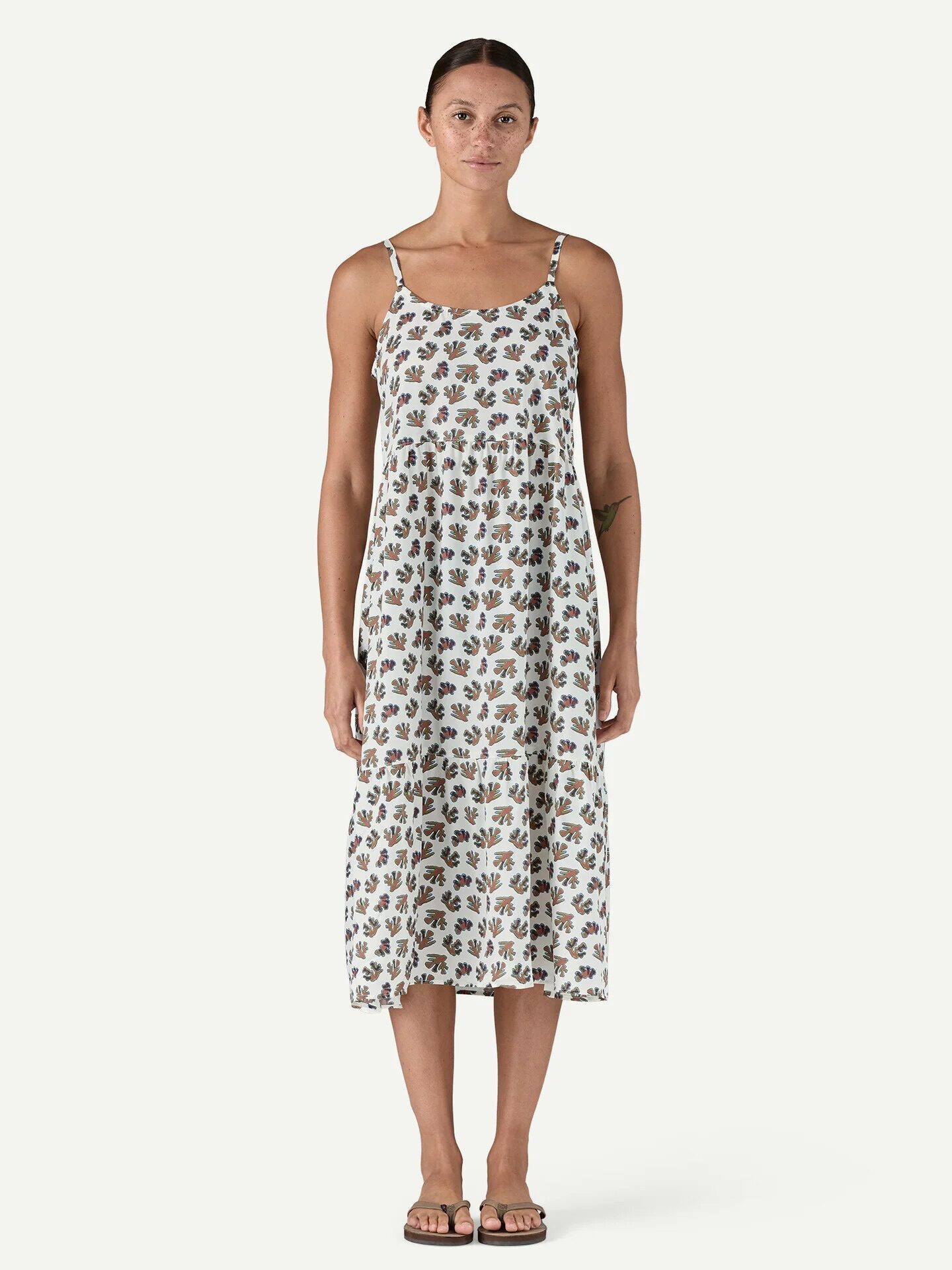 A person stands facing forward wearing a white, sleeveless, ankle-length dress with a floral print and sandals against a plain background.