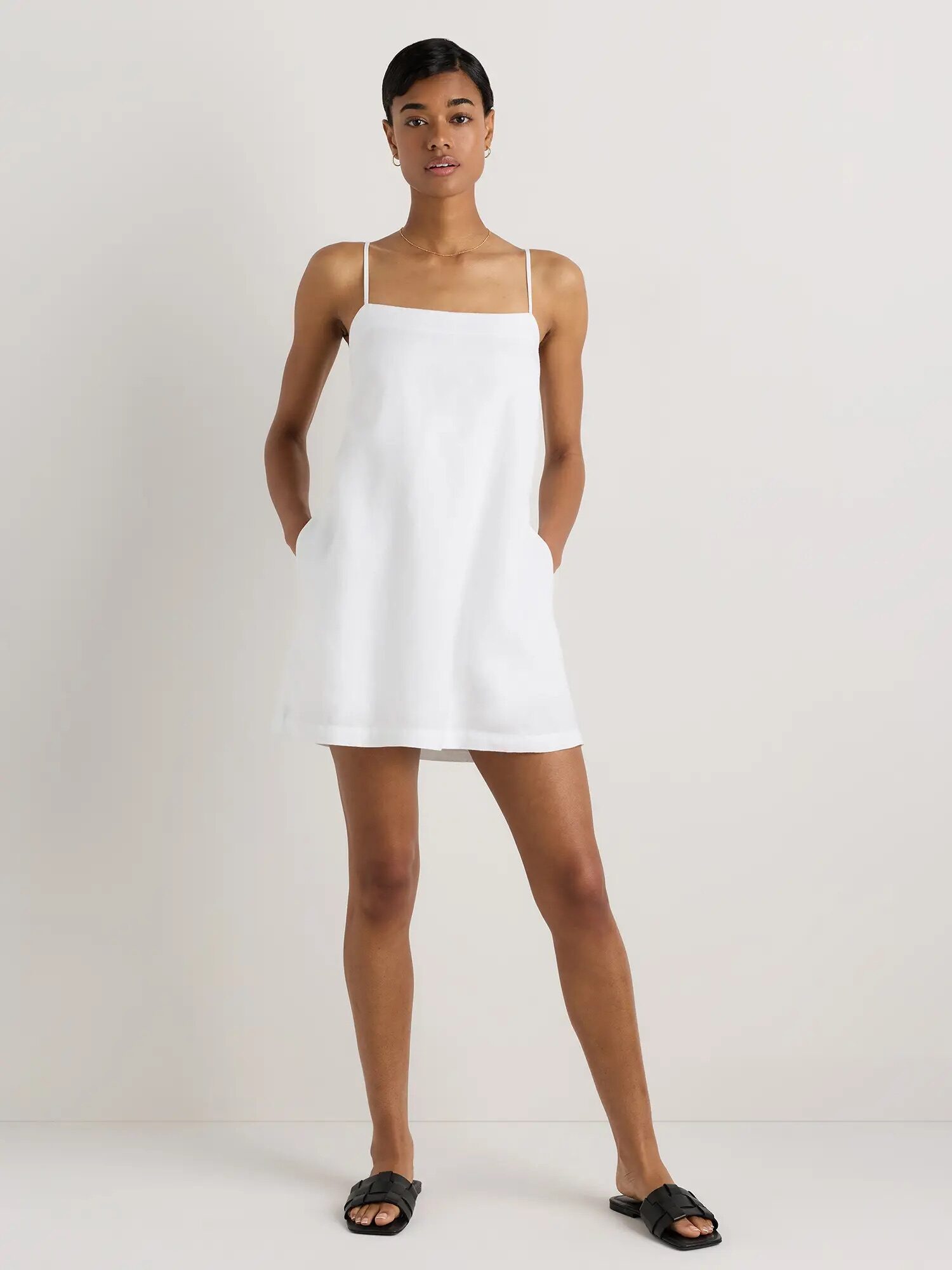 A person stands against a plain background wearing a sleeveless, white, square-neck mini dress and black sandals.