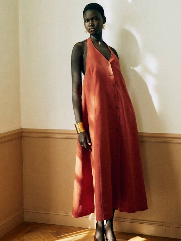 A woman stands indoors wearing a loose, sleeveless, rust-colored dress with buttons down the front, gold hoop earrings, and a gold bracelet.