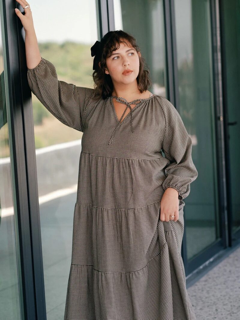 A woman in a long, checkered dress stands indoors by a large glass window, resting one hand on the frame and looking off to the side.