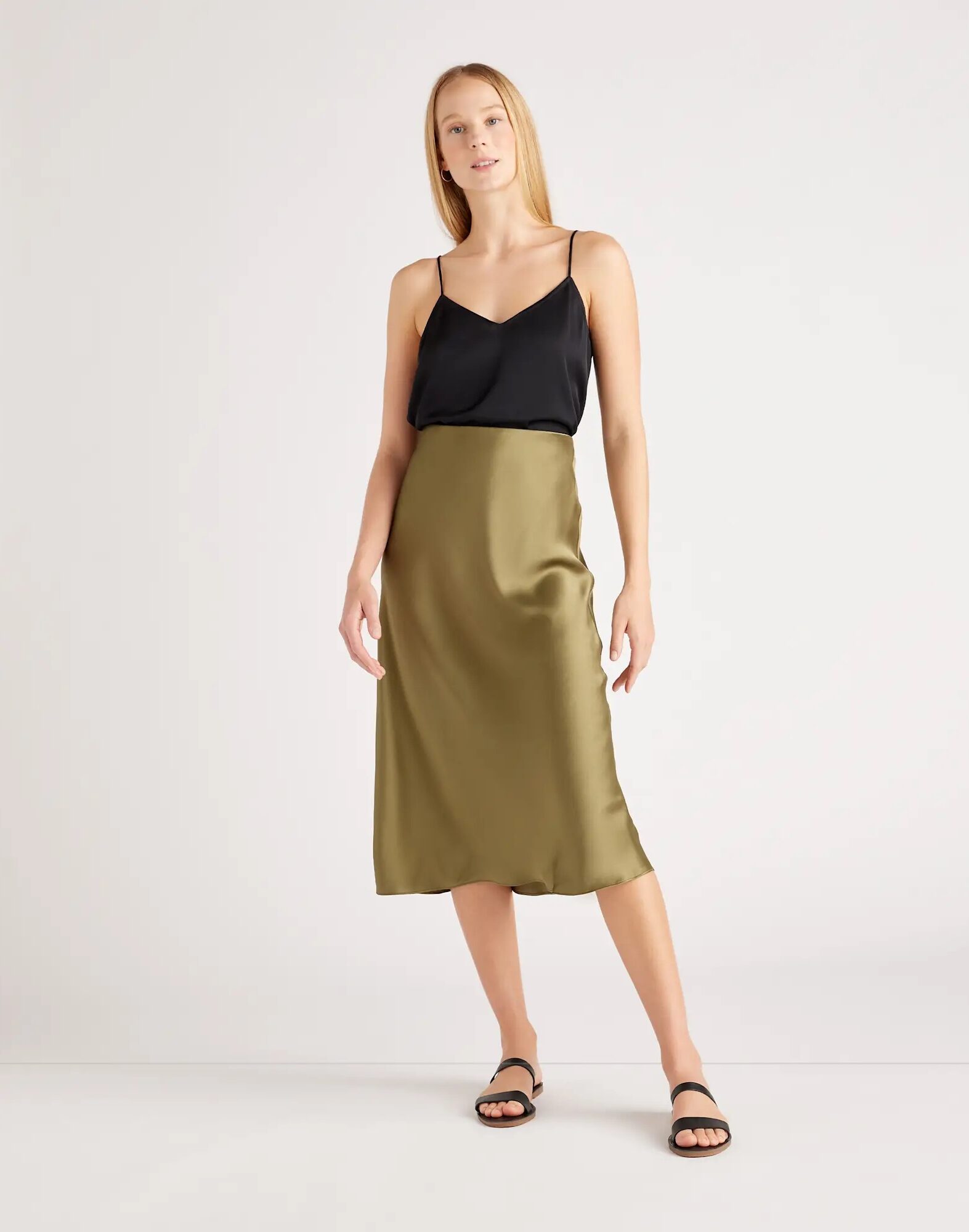 A person stands against a plain background wearing a black camisole top, an olive green satin midi skirt, and black sandals.