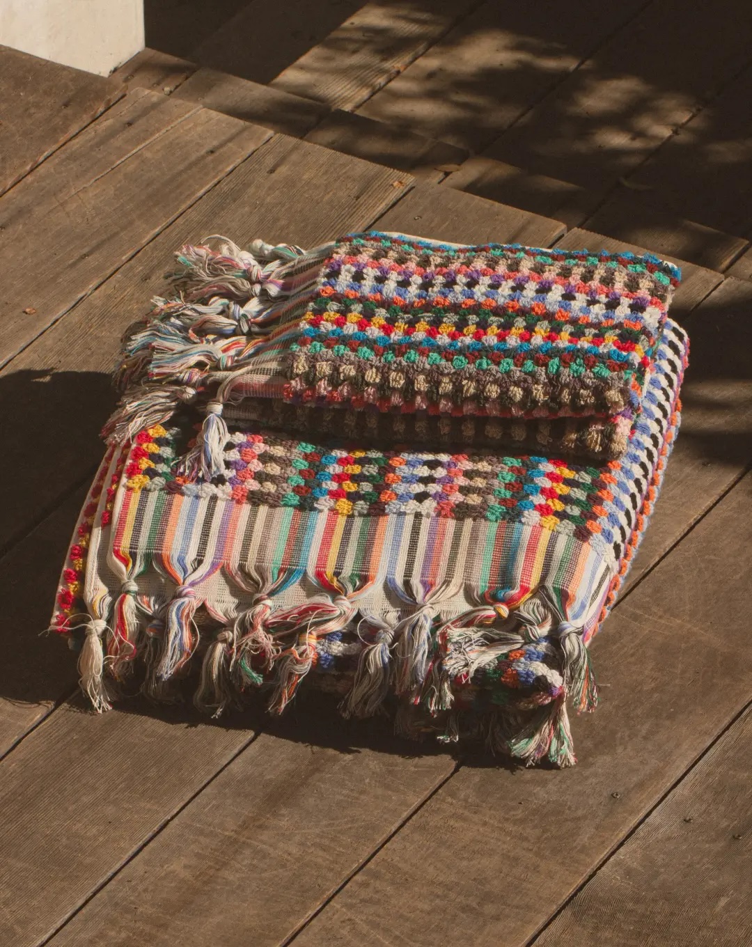 Two folded, multicolored woven blankets with fringed edges sit stacked on a wooden floor in sunlight.