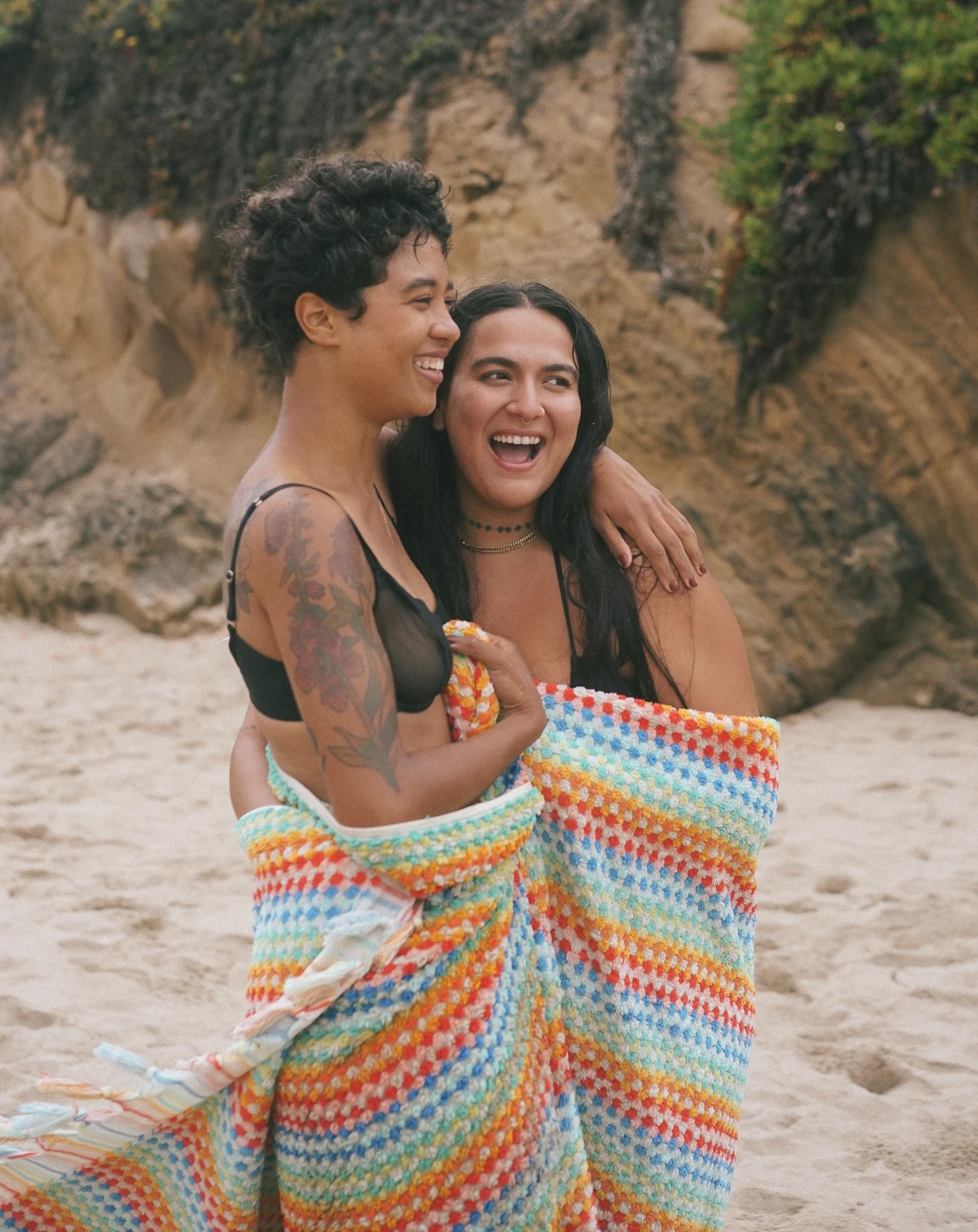 Two women stand on a sandy beach, wrapped together in a colorful crocheted blanket, smiling and embracing in front of a rocky backdrop with greenery.