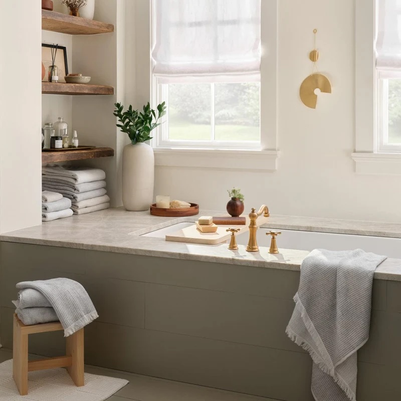 Modern bathroom with a built-in tub, gold fixtures, folded towels, a wooden stool with a gray towel, and shelves holding toiletries and decor items near two windows with white curtains.