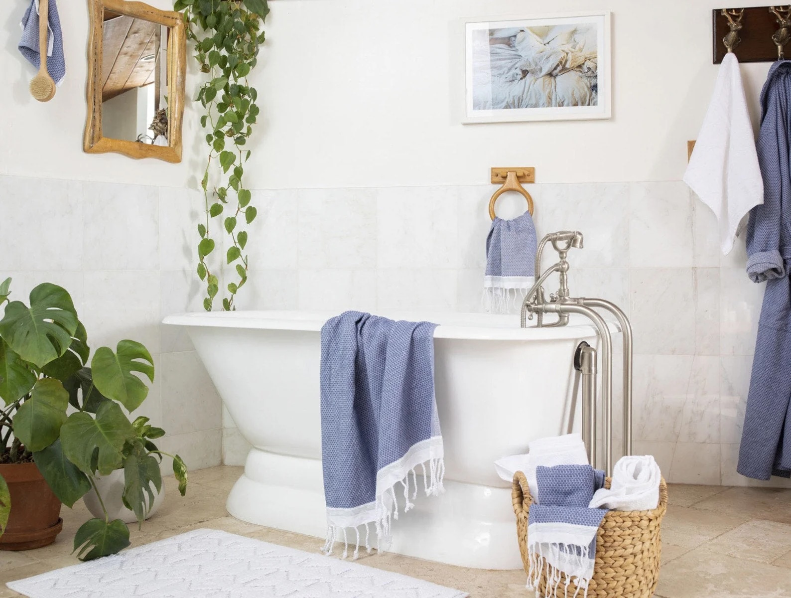 A white freestanding bathtub with blue towels, surrounded by green plants, a basket of towels, and wall-mounted art in a bright bathroom.