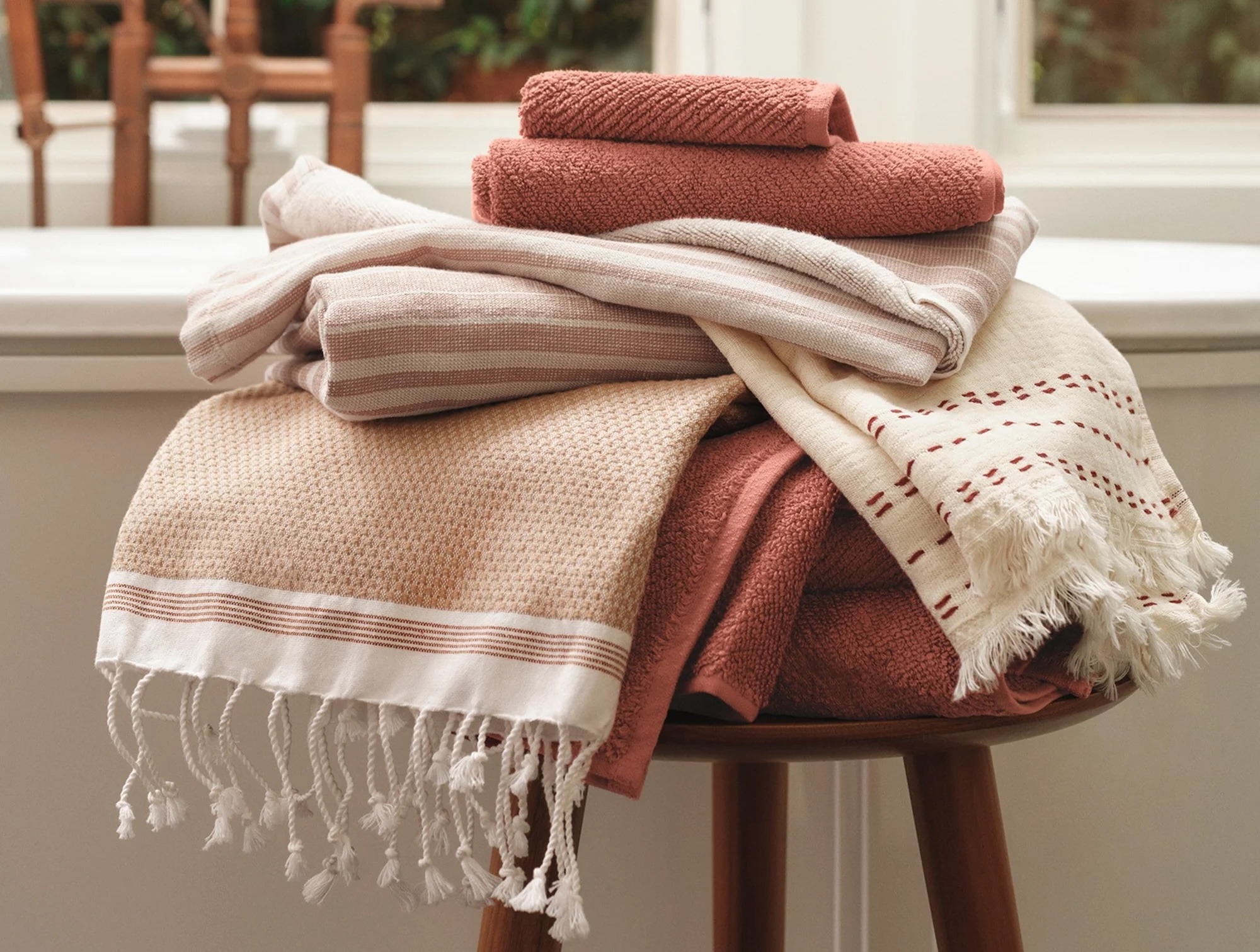 A stack of folded towels and fringed blankets in neutral and pink tones sits on a wooden stool in front of a window.