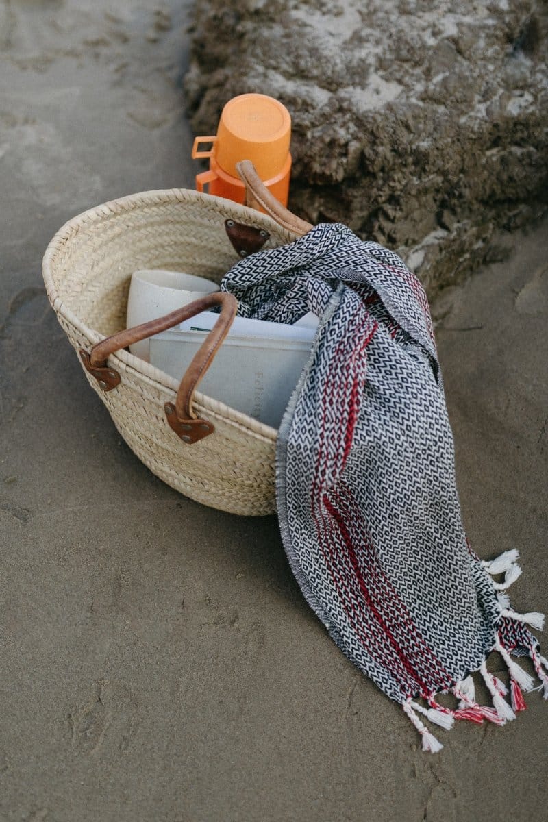 A woven beach bag with leather handles contains a towel and a magazine, sitting on sand near a rock and an orange container in the background.
