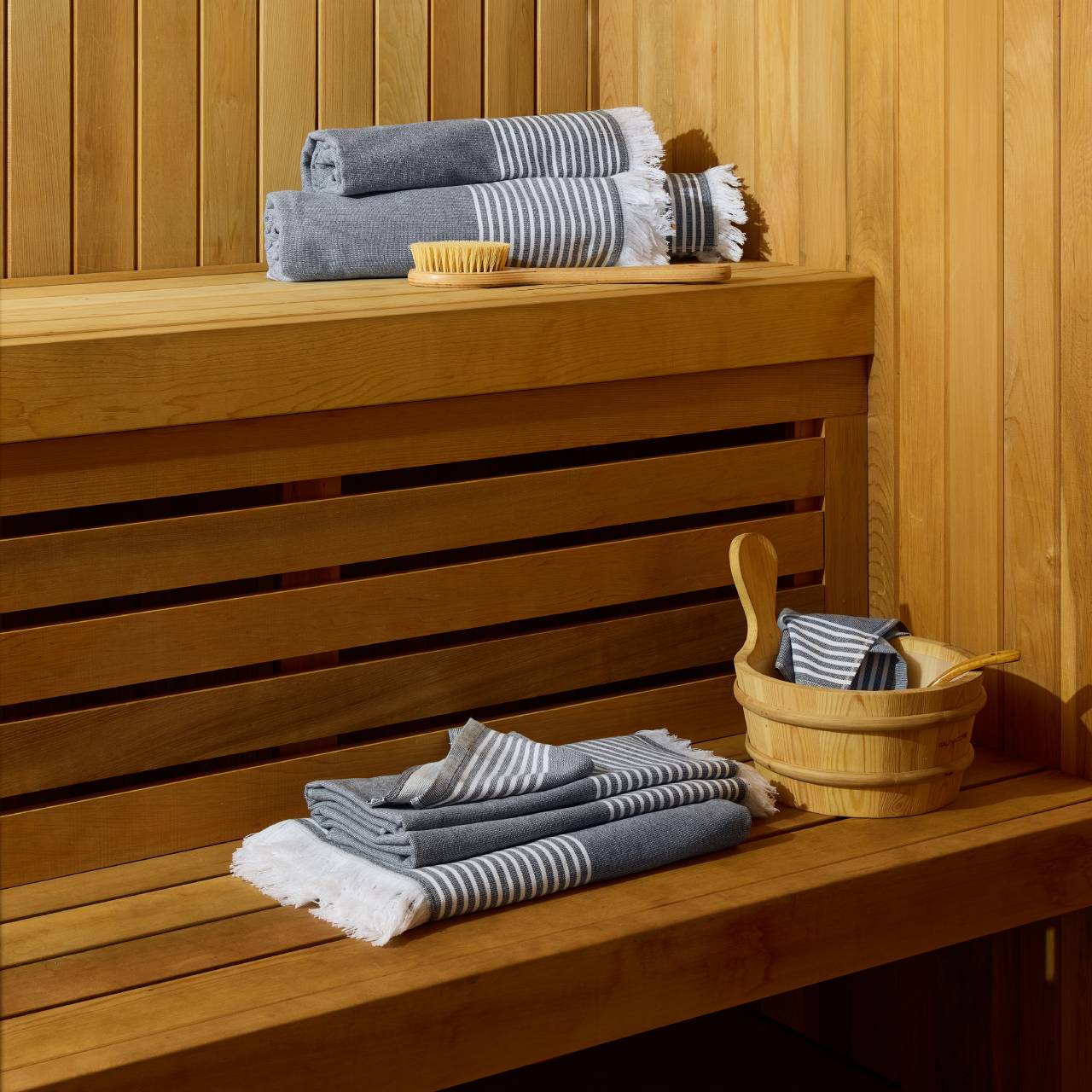 Stacked striped towels, a wooden brush, and a bucket with a ladle are arranged on wooden benches inside a sauna.