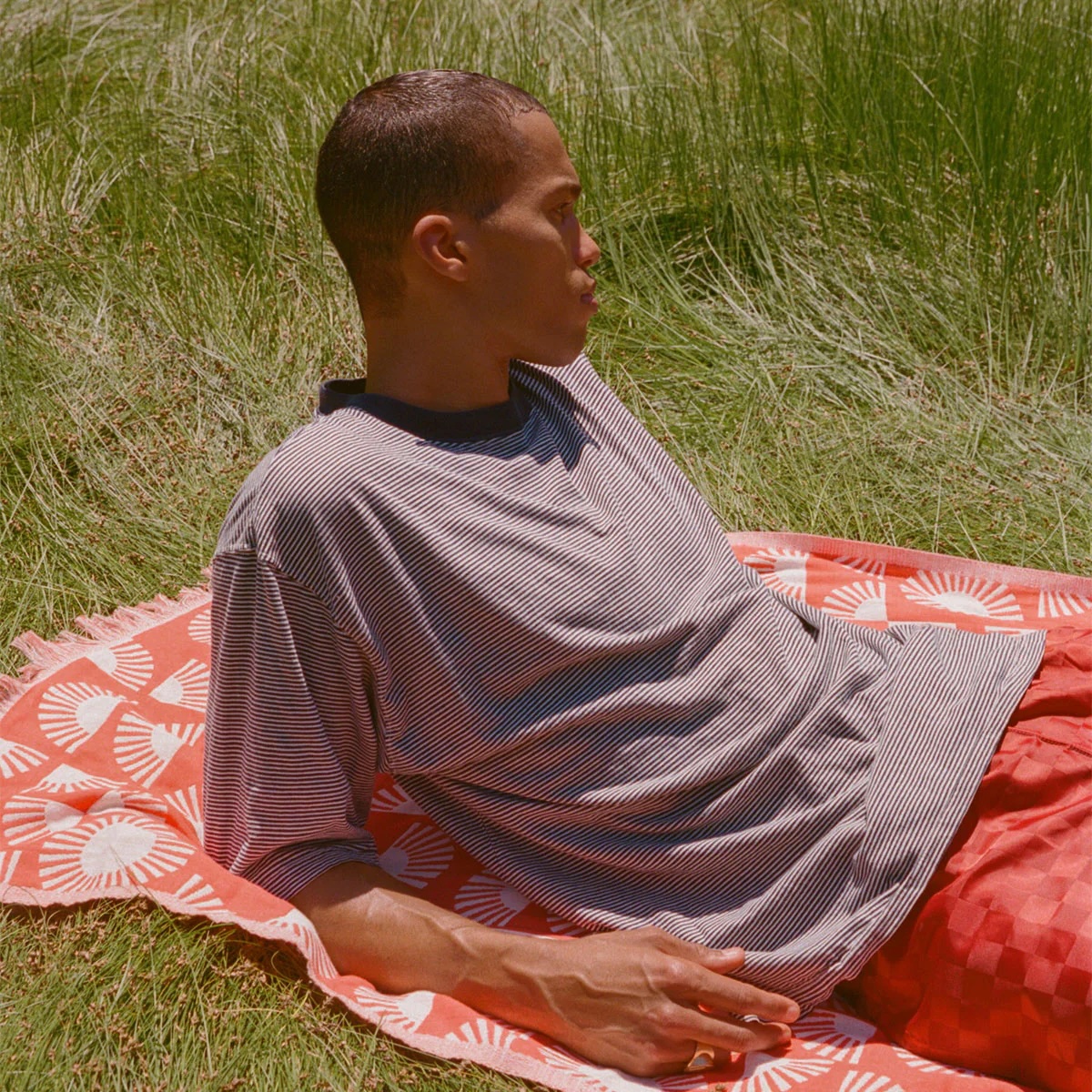 Person lying on an orange patterned blanket in the grass, wearing a striped t-shirt and red shorts, looking off to the side.
