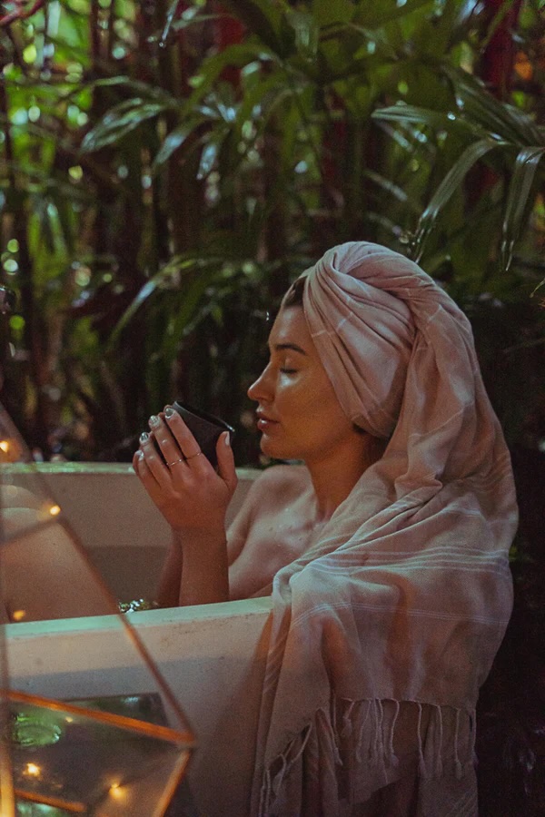 A person with a towel wrapped around their head sits in a bathtub outdoors, holding a cup, surrounded by plants and warm lighting.
