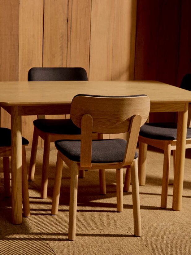 A wooden dining table with four matching chairs featuring black cushions, set in a room with wooden walls and carpeted flooring.