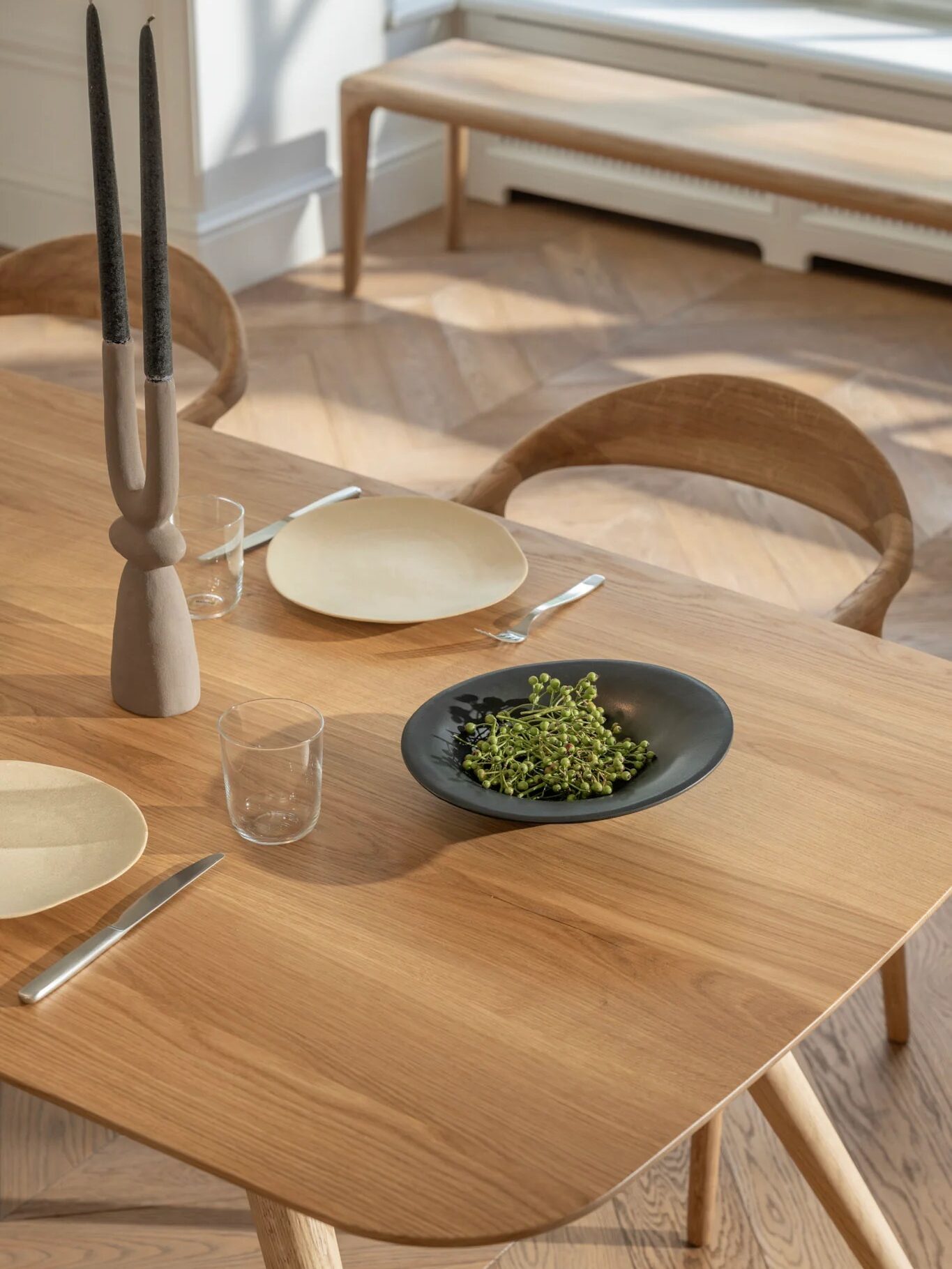 A wooden dining table with three place settings, a black bowl of green herbs, clear glasses, and a decorative candleholder. Wooden chairs and a bench are in the background.