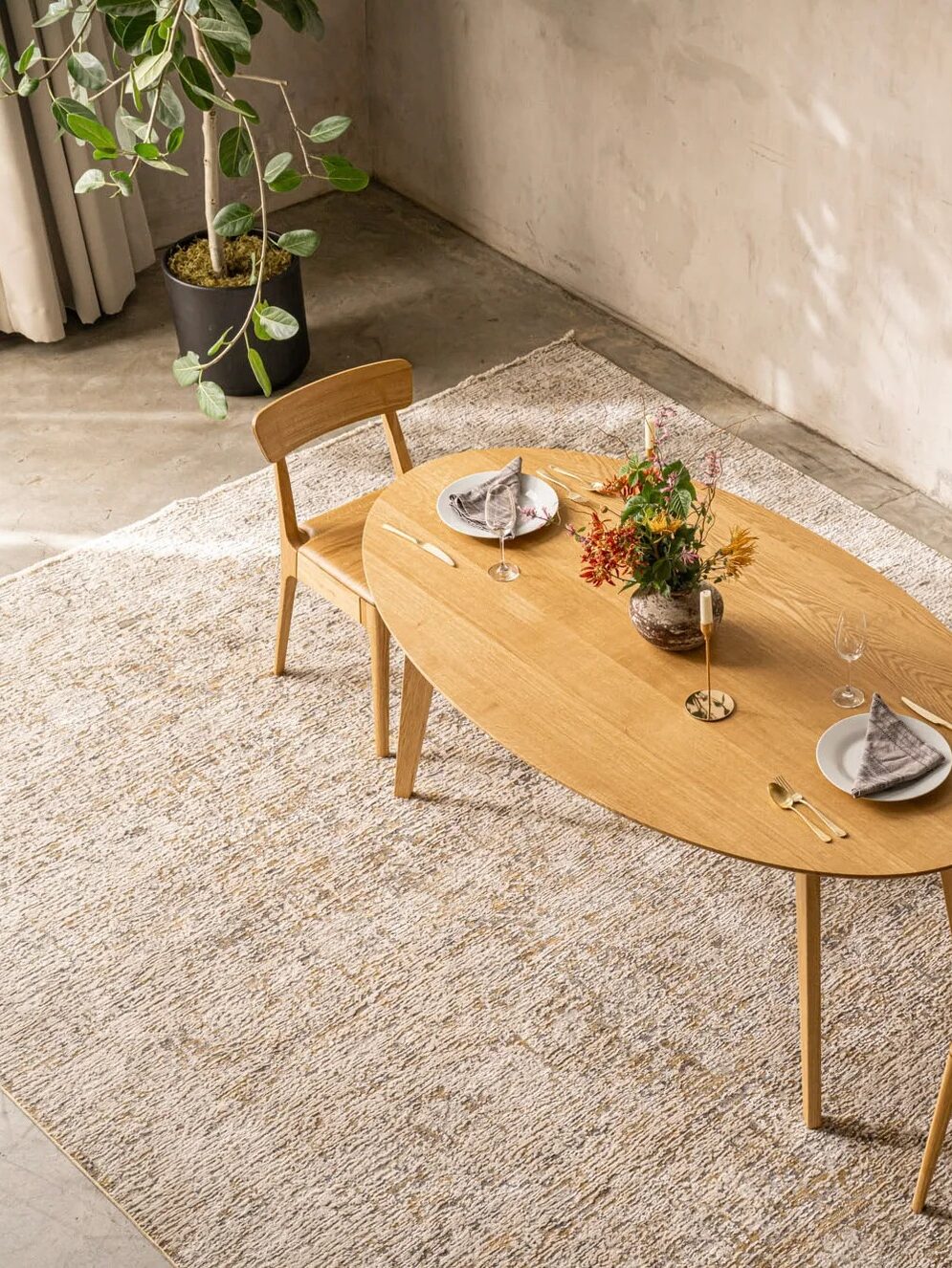 Oval wooden dining table with two chairs, set for two with plates, glasses, and napkins, centerpiece with flowers, on a woven rug in a minimalist room with a floor lamp and potted plant.