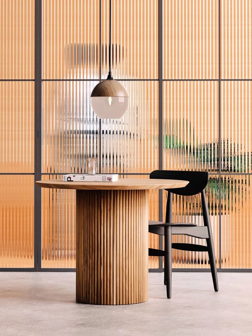 A modern round wooden table with a black chair, a book, and pendant lamp, set in front of vertical glass panels with a blurred figure and plant in the background.