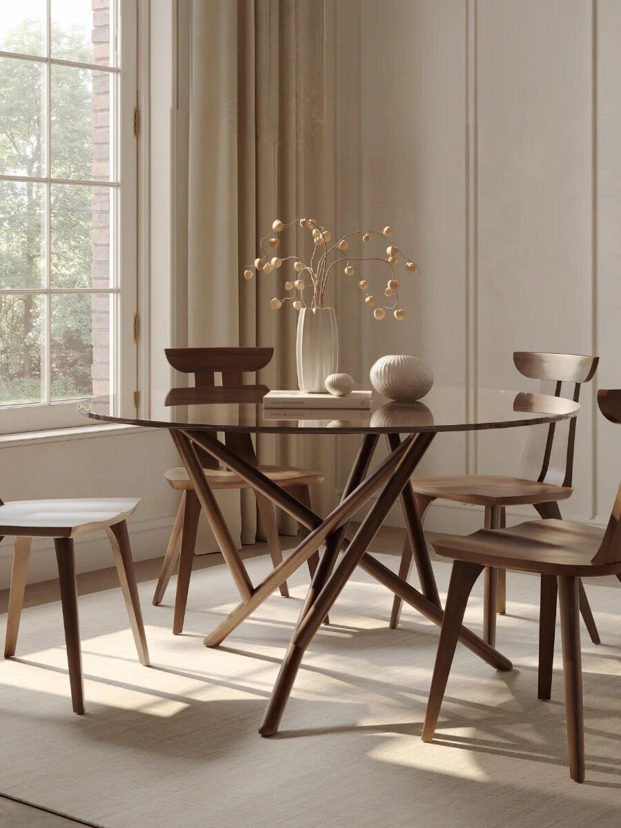 A modern dining room with a round glass table, four wooden chairs, decorative vases, and dried branches, set near large windows with beige curtains.