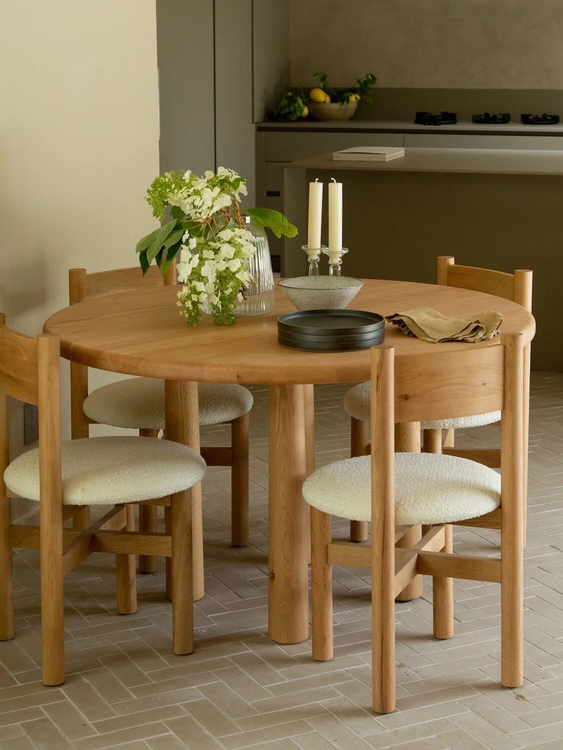 A round wooden dining table with four chairs is set with a flower vase, candles, and dishes in a minimalist kitchen with neutral tones and a bar stool in the background.