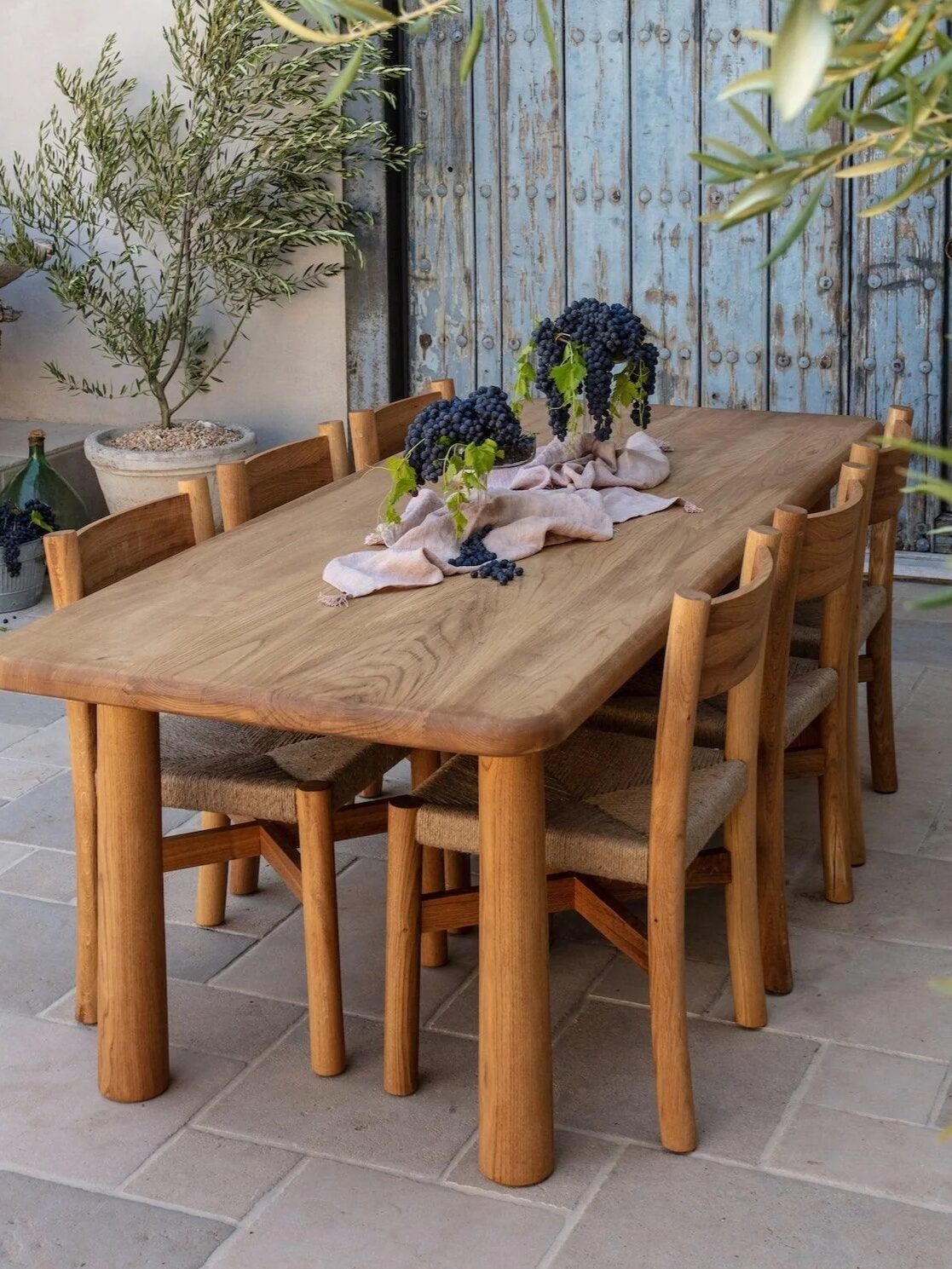 A wooden dining table with six chairs is set outdoors on stone tiles, decorated with bunches of grapes and linen runners; potted plants and a rustic blue door are in the background.