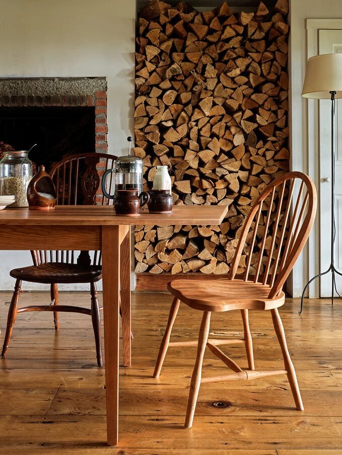 A wooden dining table and chairs sit on a wooden floor, with a large stacked pile of firewood against the wall in the background.