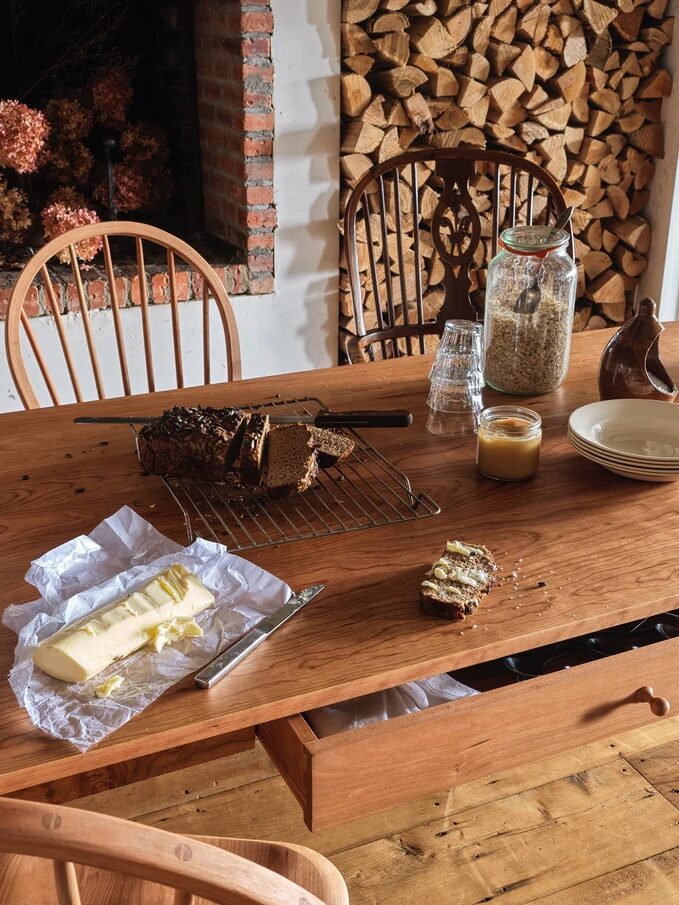 A wooden table with sliced bread on a cooling rack, butter on paper with a knife, a jar of oats, a honey jar, stacked plates, and two wooden chairs; firewood stacked in the background.