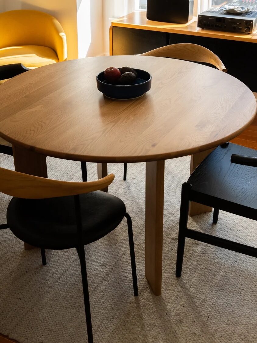 A round wooden dining table with four chairs, one with a light wood backrest and the others dark, sits on a light rug; closeup shows table edge and chair detail.