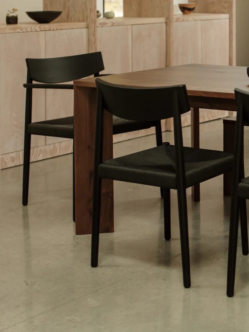 A modern dining area with a wooden table, four black chairs, and tabletop items including a bowl, plate of oranges, and a metal carafe on a polished concrete floor.