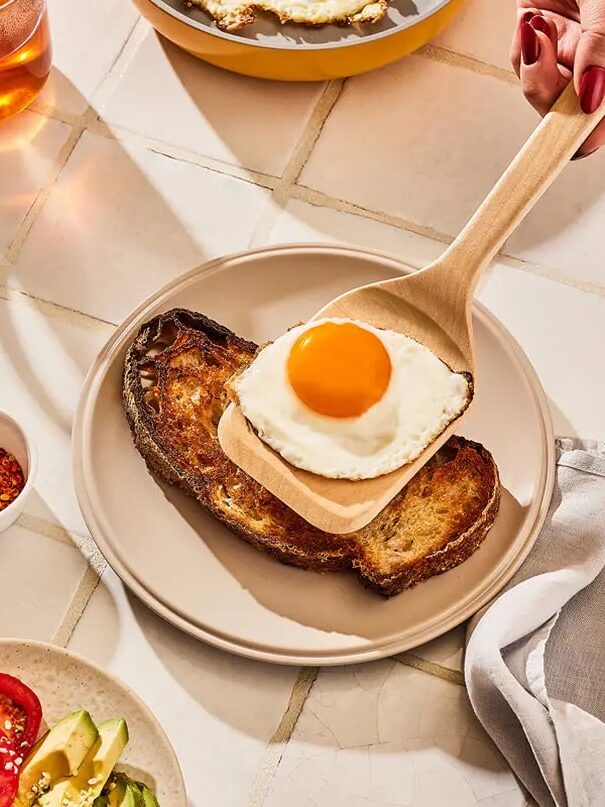 A hand places a sunny-side-up egg onto a slice of toast on a plate. Nearby are a cup of tea, a bowl of chili flakes, and a plate with sliced tomato and avocado.