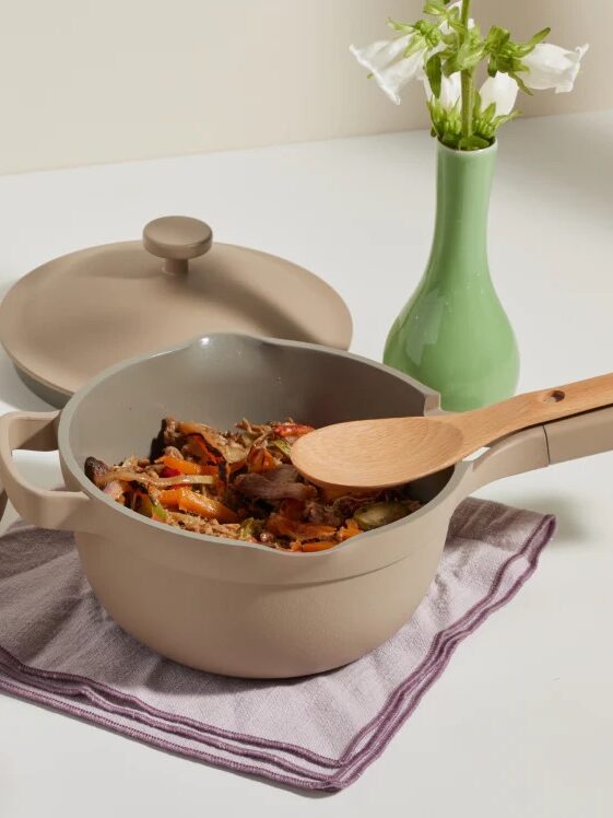 A beige pot with a wooden spoon rests on a folded cloth, filled with cooked vegetables, next to a green vase with white flowers on a white surface.
