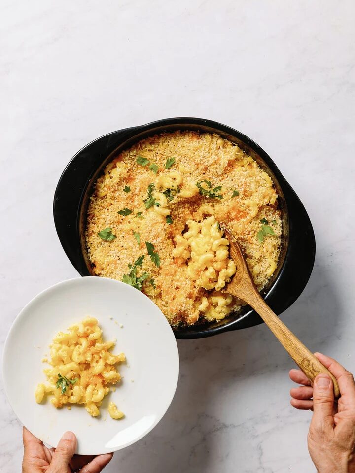A person serving baked macaroni and cheese with breadcrumbs from a black casserole dish onto a white plate.
