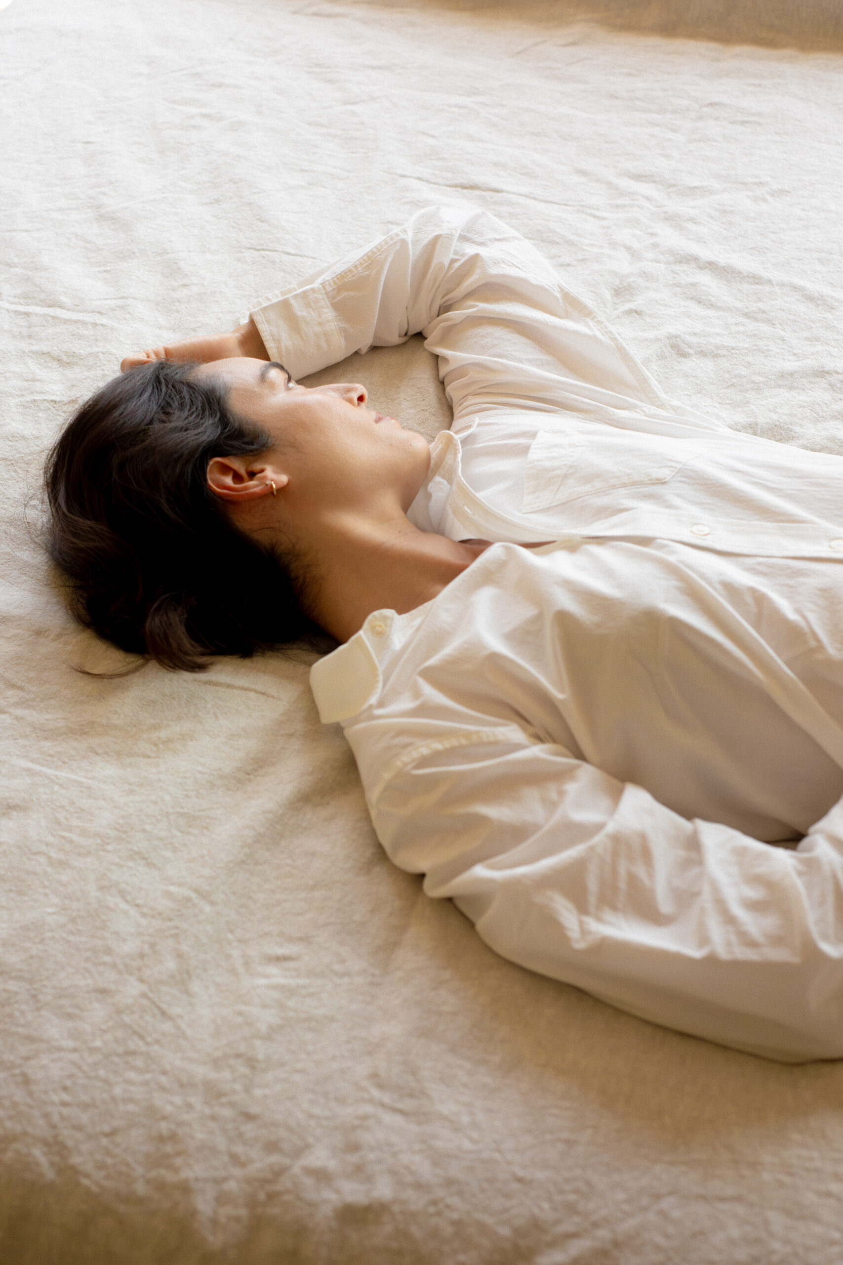 Person in a white shirt lying on their side on a light-colored bed, with one arm bent and resting near their head.