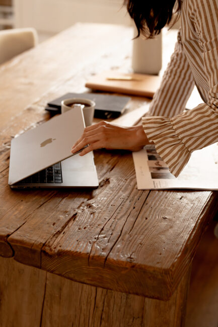 A person wearing a striped shirt closes a laptop on a rustic wooden table with papers, a coffee cup, and electronic devices nearby.