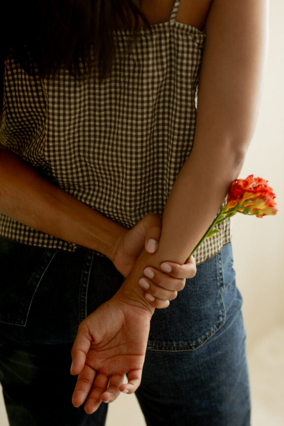 A person in a checkered top and jeans holds an orange carnation behind their back with both hands.