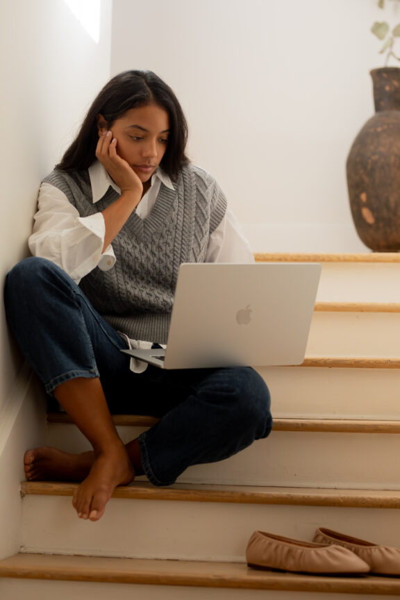 A woman sits barefoot on stairs, leaning on one hand and looking at a laptop screen. A bag lies on a lower step and a large decorative vase is in the background.