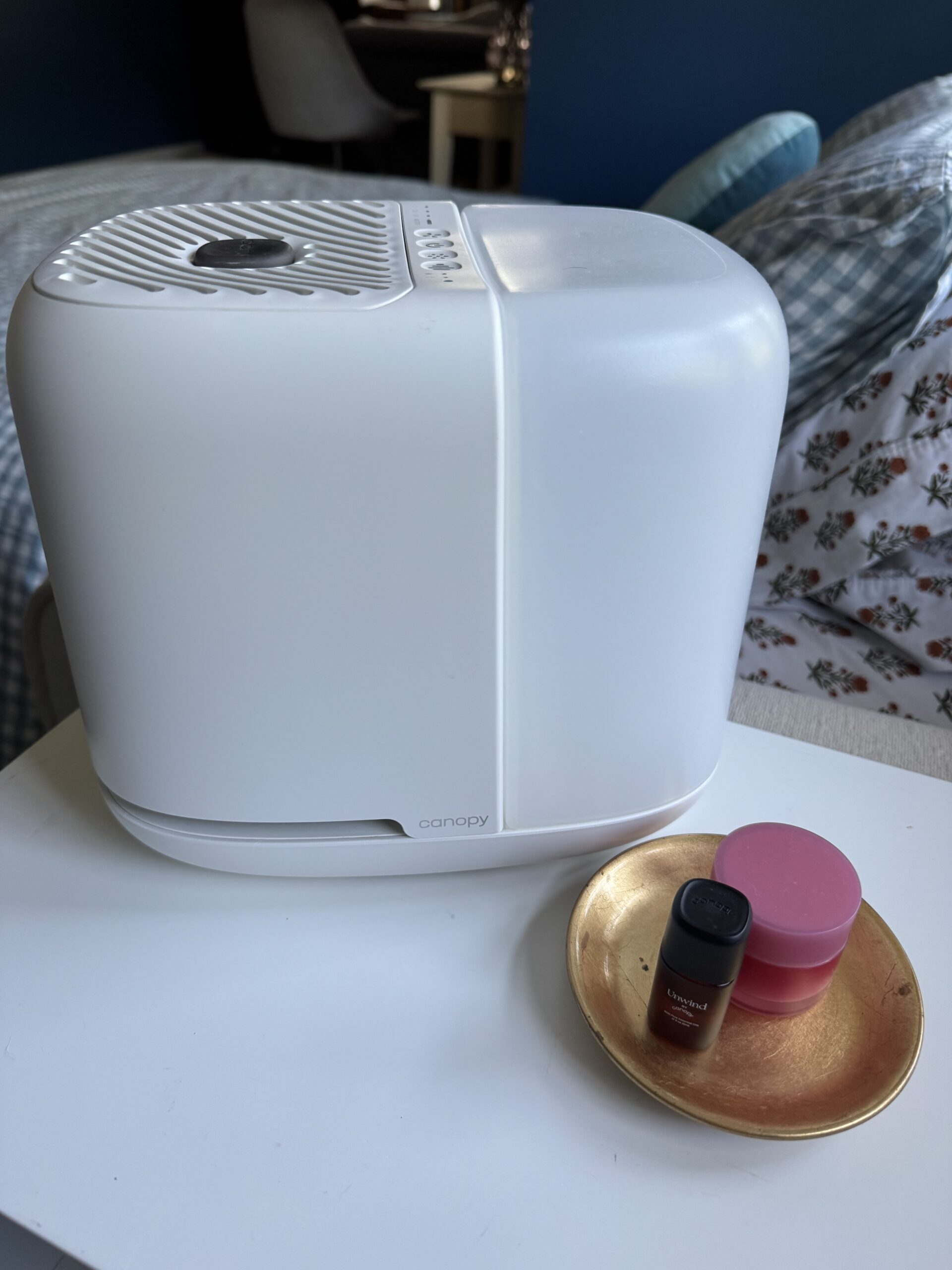 A white Canopy humidifier sits on a white surface next to a small gold tray holding a dark bottle and a pink container. A bed and chair are visible in the background.