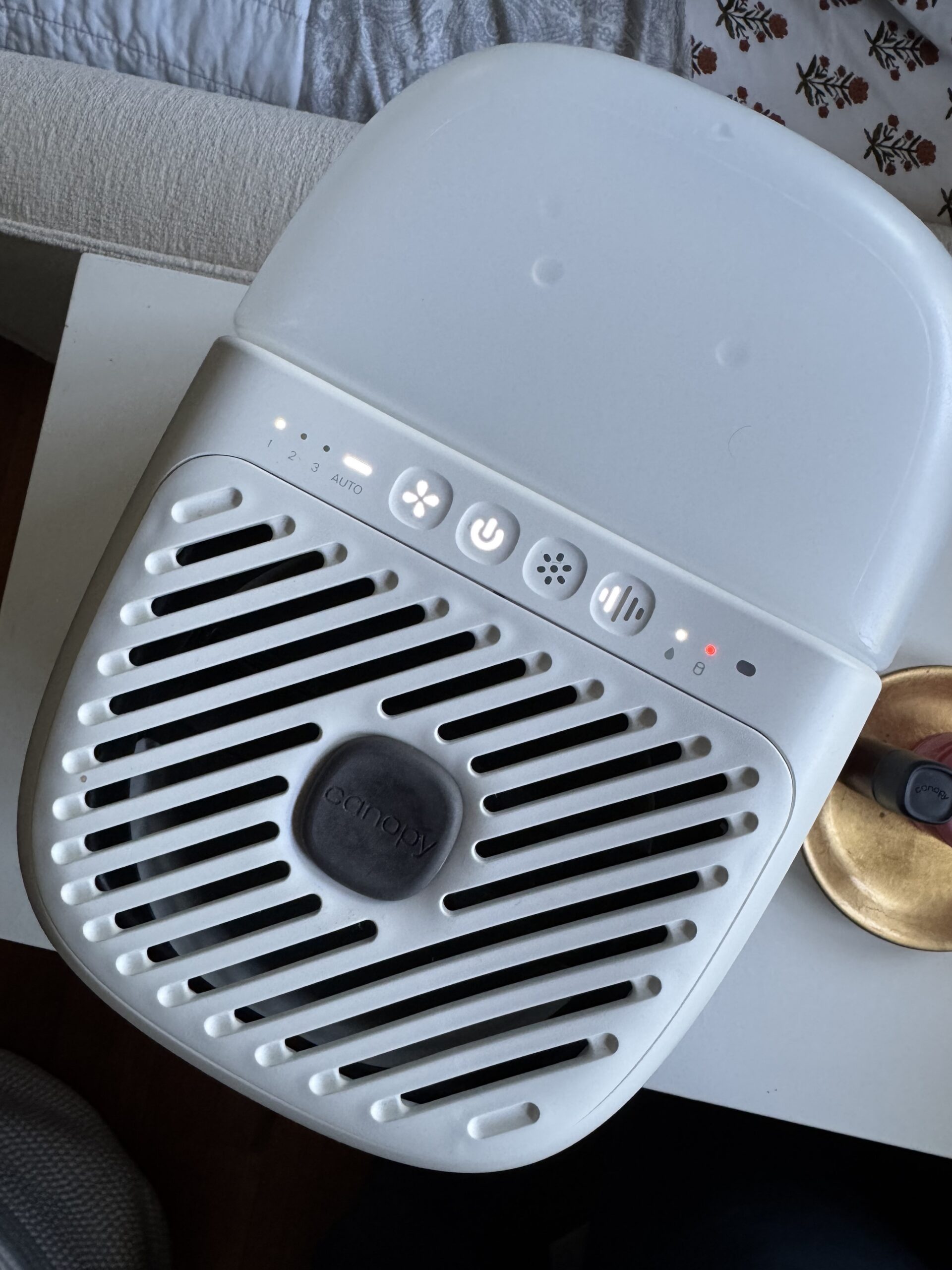 A white air purifier with control buttons and indicator lights is placed on a bedside table next to a gold dish and part of a bed with a patterned blanket.