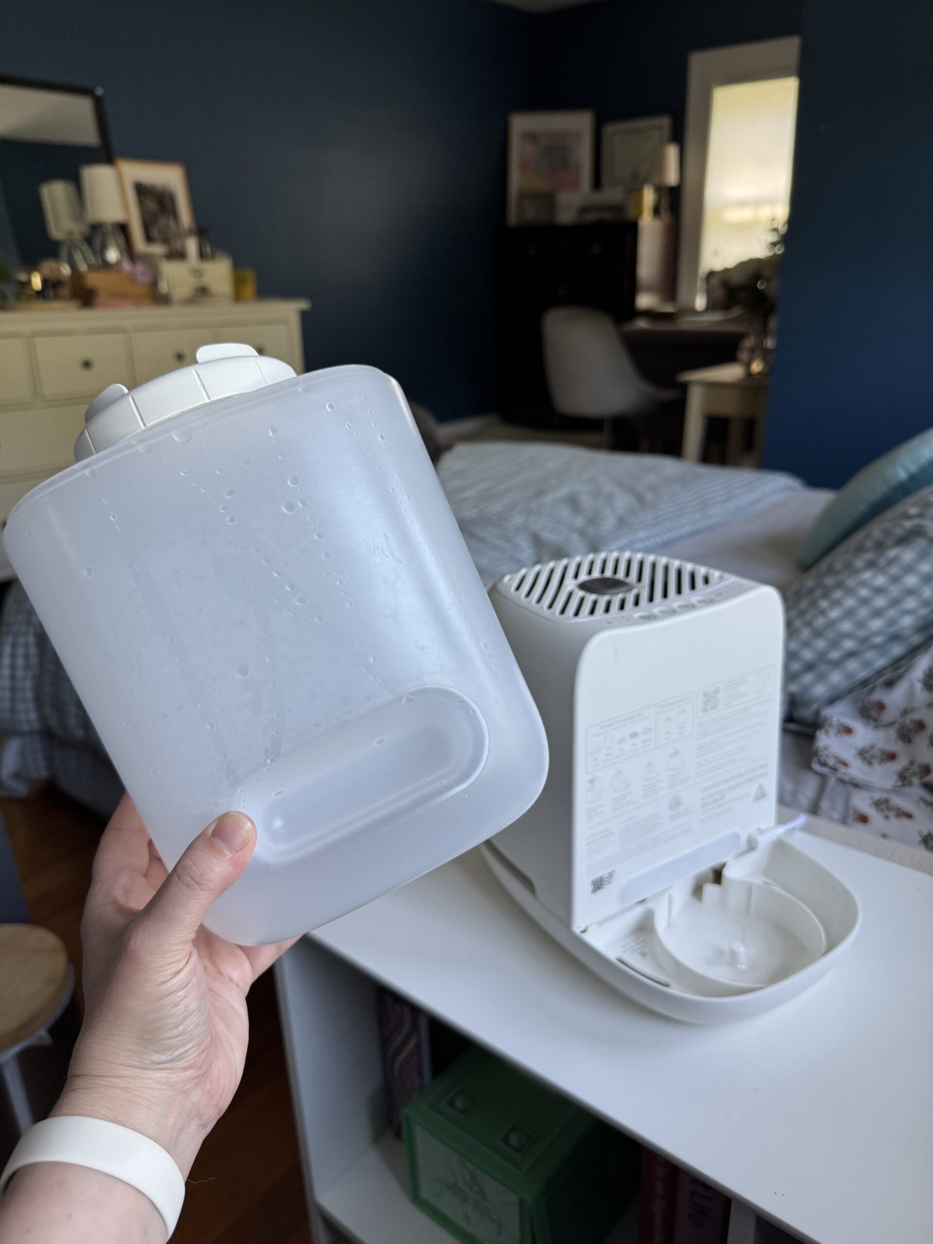 A hand holds a detached water tank in front of a white humidifier in a bedroom with blue walls and a bed in the background.