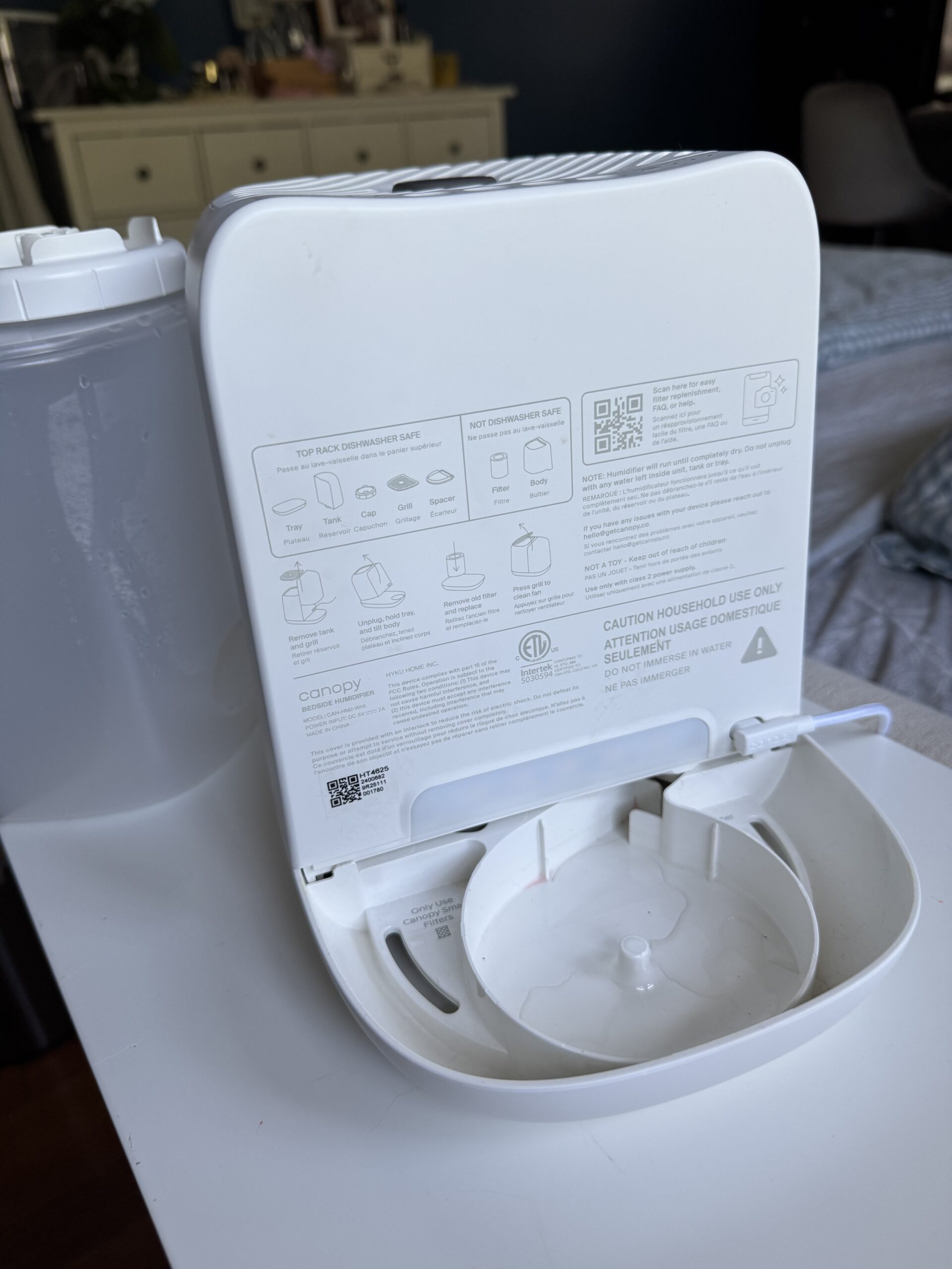 A white portable humidifier with its water tank removed, showing the open base and instruction labels on the inside panel. The unit sits on a white surface in a bedroom.