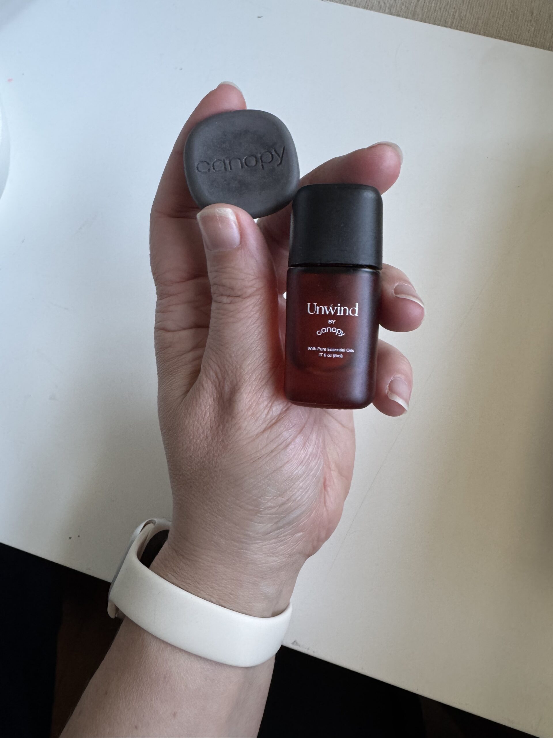 A hand holding a small dark bottle labeled "Unwind" and a black disk labeled "canopy," with a white wristband visible on the wrist.