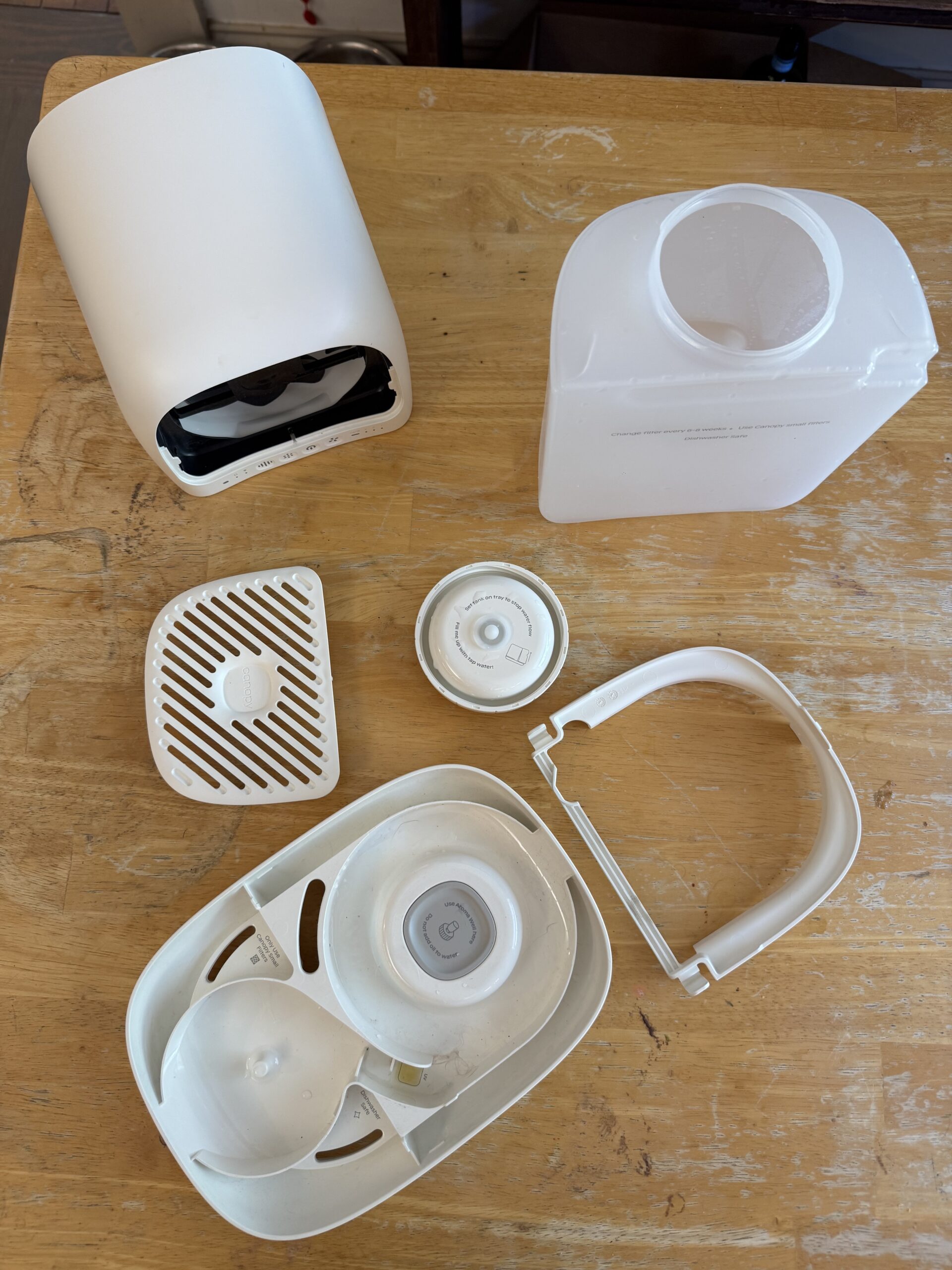 Disassembled white humidifier with its water tank, filter, lid, handle, and base components spread out on a wooden surface.